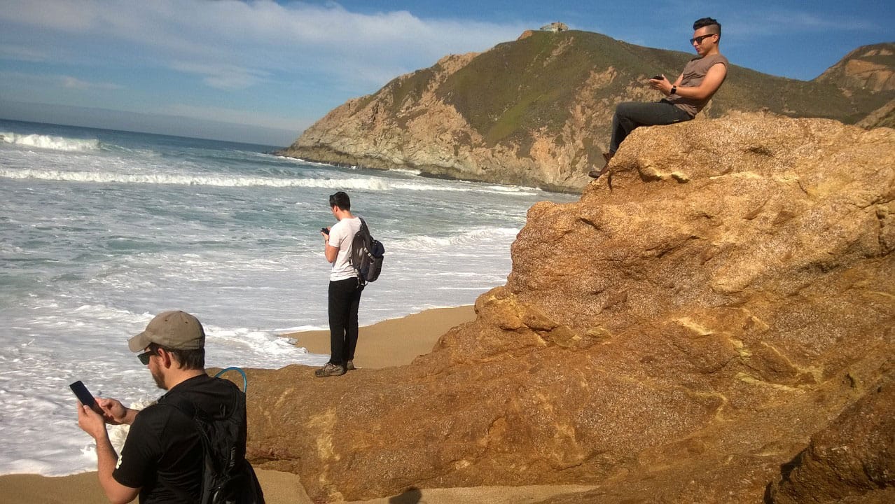 Three men on the ocean shore looking at their phones.