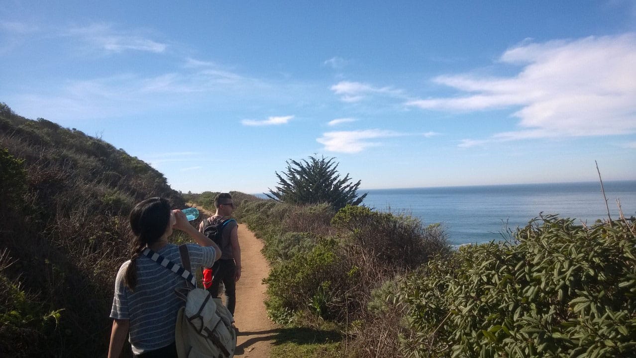 Two figures walking along a mountain trail with the ocean in the background.